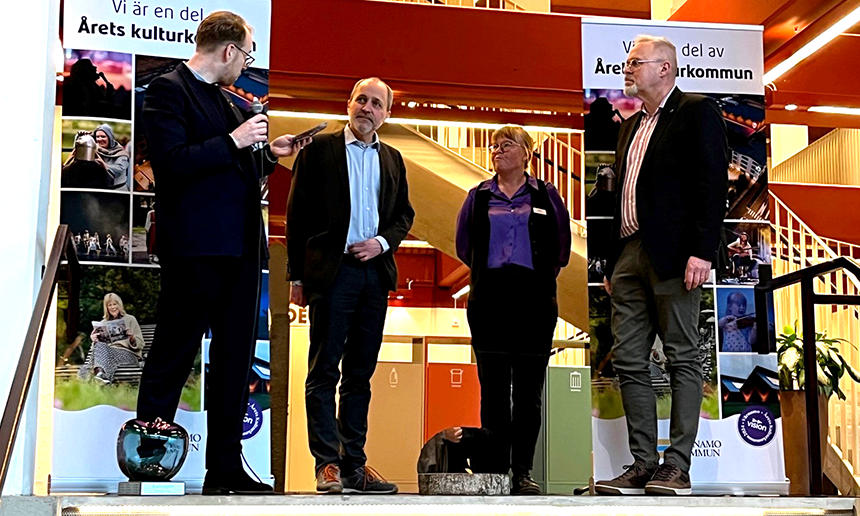 Fyra personer står på ett podium, en av dem håller i en mikrofon. Längst fram till vänster står ett äpple i glas.