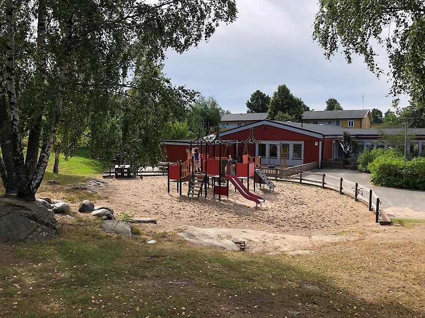 En bild från Trädkryparens gård. Det finns en rutschkana och en klättervägg. Förskolans hus syns i bakgrunden. Det är en röd byggnad med grå knutar